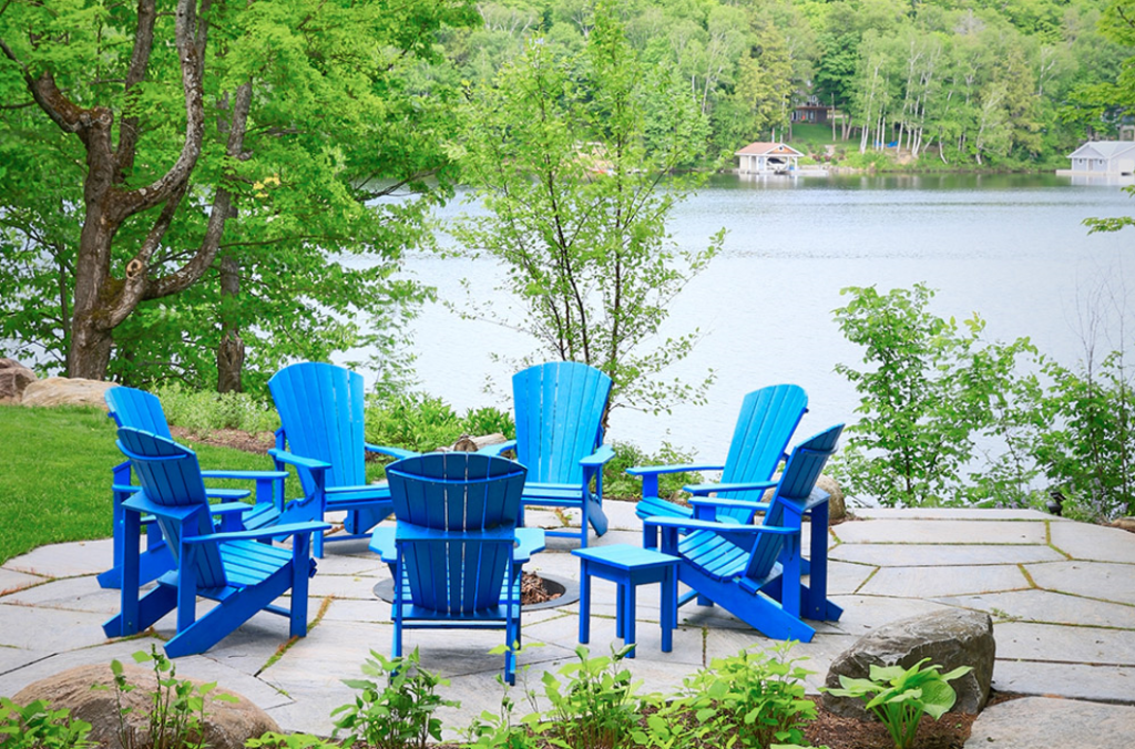 7 Blue Adirondack chairs surrounding a fireplace next to a pond.