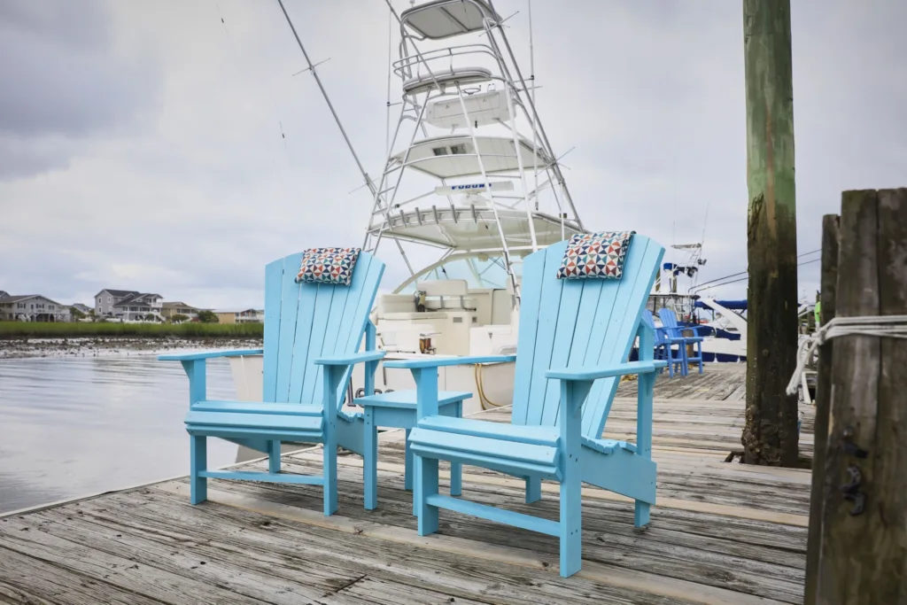 Two sky blue Generations Upright Adirondack Chairs on a dock next to the ocean, with a sailboat in the background